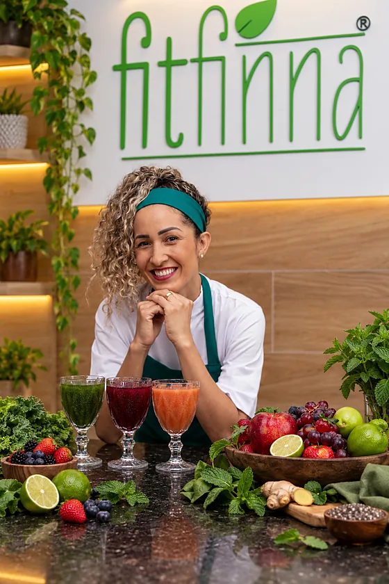 Zélia Gomes sorrindo, de avental Fitfinna, atrás de três sucos naturais e uma tigela de frutas, com a marca ao fundo.